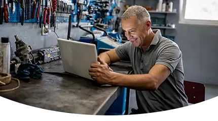 A smiling mechanic uses a laptop at his workbench in front of a pegboard of tools.